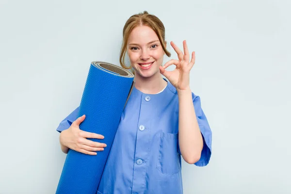 年轻的高加索理疗师 拿着一块被蓝色背景隔离的垫子 快乐而自信地做出了好的姿势 — 图库照片