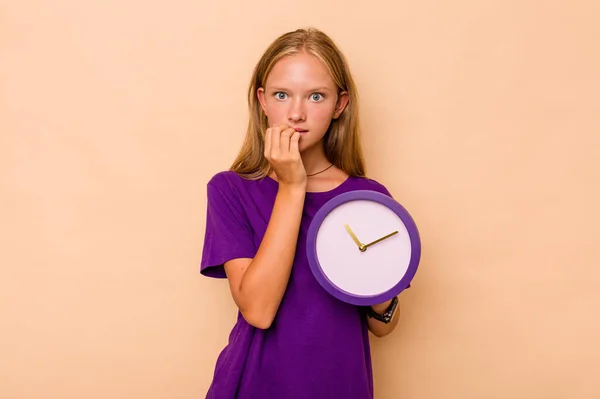 Menina Caucasiana Segurando Relógio Isolado Fundo Bege Mordendo Unhas Nervoso — Fotografia de Stock