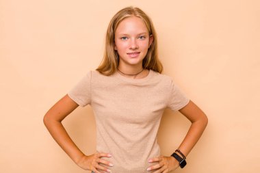 Beyaz tenli genç kız, bej arka planda izole edilmiş. Kendine güvenen, ellerini kalçasında tutan..