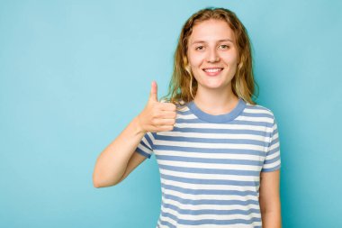 Mavi arka planda izole edilmiş genç beyaz kadın gülümsüyor ve başparmağını kaldırıyor.