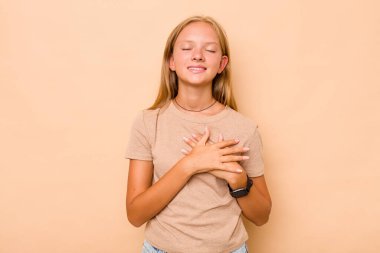 Beyaz tenli genç kız bej arka planda izole edilmiş. Gülüyor, elini kalbe koyuyor, mutluluk kavramını koruyor..
