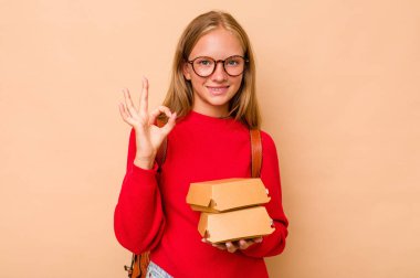 Elinde hamburgerler tutan küçük beyaz kız bej arka planda izole edilmiş neşeli ve kendinden emin..