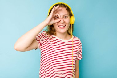 Mavi arka planda izole edilmiş kulaklık takan beyaz bir kadın heyecanlı..