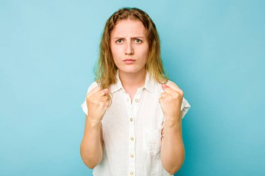 Mavi arka planda izole edilmiş genç beyaz kadın yumruğunu kameraya gösteriyor, agresif yüz ifadesi..