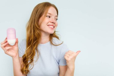 Elinde deodorant tutan beyaz bir kadın mavi arka planda izole edilmiş, baş parmak uzakta, gülen ve kaygısız..