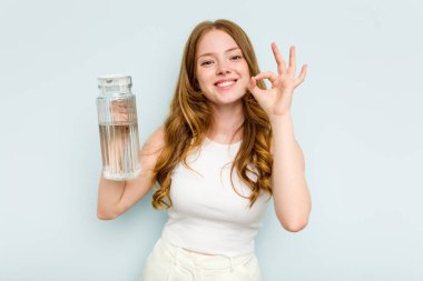 Beyaz bir kadın elinde bir kavanoz su tutarken mavi arka planda izole edilmiş neşeli ve kendinden emin..