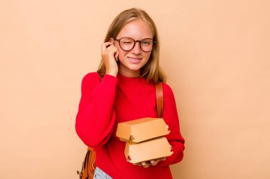 Elinde hamburgerler tutan küçük beyaz kız bej arka planda izole edilmiş kulakları elleriyle kapatıyor..