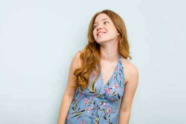 Genç beyaz kadın mavi arka planda izole edilmiş rahat ve mutlu bir şekilde gülüyor, boynunda dişlerini gösteriyor..