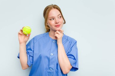 Beyaz tenli genç fizyoterapist elinde mavi arka planda izole edilmiş bir tenis topu tutuyor. Şüpheli ve şüpheci bir ifadeyle yan yana bakıyor..