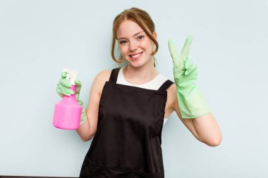 Mavi arka planda izole edilmiş genç temizlikçi kadın iki numarayı parmaklarıyla gösteriyor..