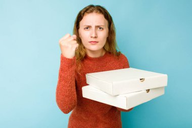 Mavi arka planda izole edilmiş pizza kutuları tutan beyaz bir kadın kameraya yumruk sallıyor, agresif bir yüz ifadesi var..
