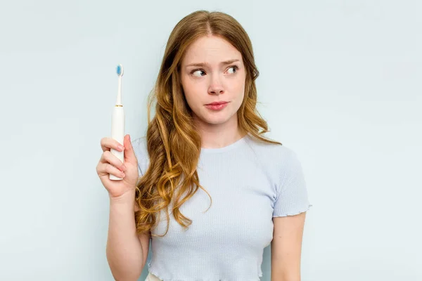 年轻的高加索女人拿着被蓝色背景隔离的电动牙刷 感到困惑 怀疑和不确定 — 图库照片