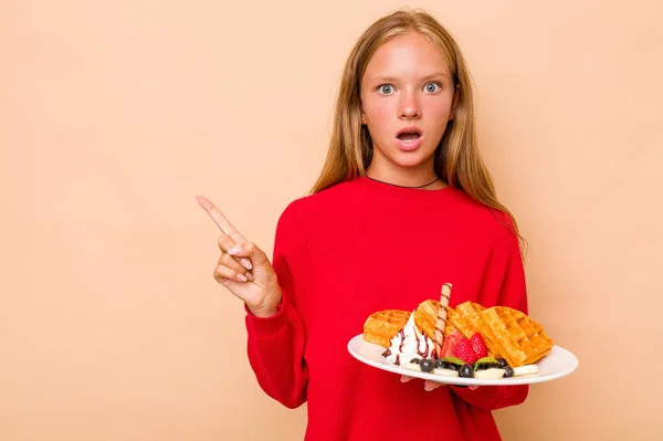 一个小高加索女孩拿着一个因米色背景而被隔离的华夫饼 指着侧面 — 图库照片