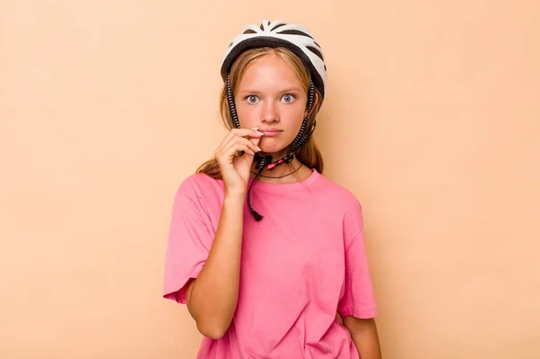 一个带着自行车头盔的高加索小女孩 带着米黄色背景的隔离帽 手指放在嘴唇上 保守秘密 — 图库照片