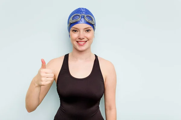 年轻的高加索游泳女子 被蓝色背景隔离 面带微笑 竖起大拇指 — 图库照片