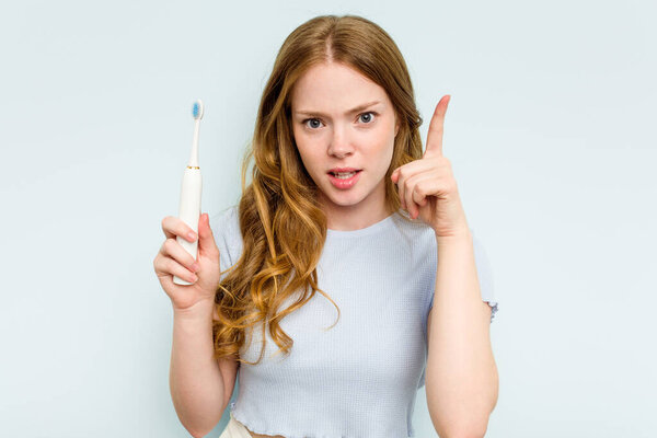 Young caucasian woman holding electric toothbrush isolated on blue background having an idea, inspiration concept.