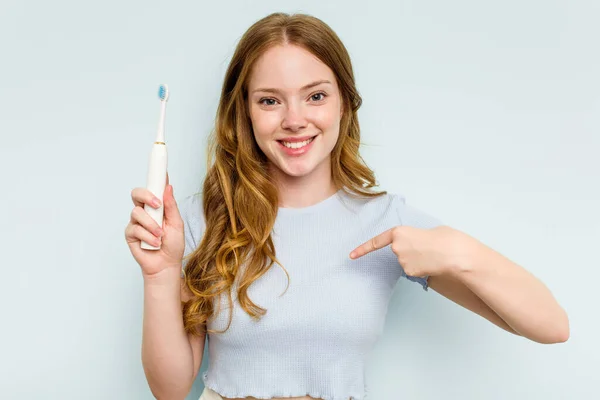 青い背景の人に手でシャツのコピースペースを指して隔離された電動歯ブラシを保持若い白人女性 誇りと自信 — ストック写真