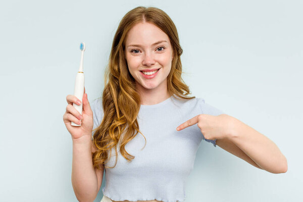 Young caucasian woman holding electric toothbrush isolated on blue background person pointing by hand to a shirt copy space, proud and confident