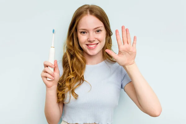 年轻的高加索女人拿着被蓝色背景隔离的电动牙刷 高兴地微笑着 用手指显示出5号 — 图库照片