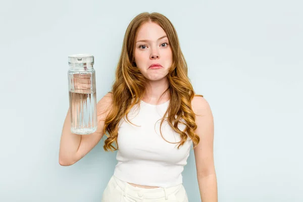 stock image Young caucasian woman holding jar of water isolated on blue background shrugs shoulders and open eyes confused.