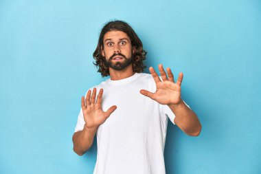 Beyaz tişörtlü sakallı adam, mavi arka plan, yakın bir tehlike nedeniyle şoka giriyor.