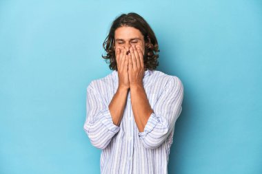 Mavi çizgili gömlekli sakallı bir adam, mavi stüdyo bir şeye gülüyor, elleri ağızda..