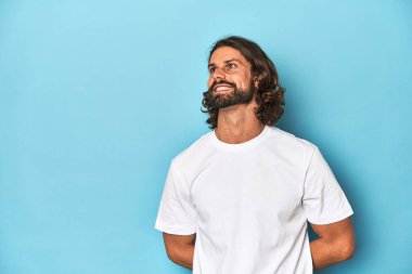 Beyaz gömlekli sakallı adam, mavi arka planda gevşemiş ve mutlu bir gülüş, dişlerini göstererek boynunu germiş..