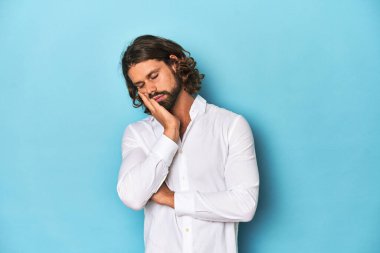 Beyaz tişörtlü sakallı adam, mavi arka plan, sıkılmış, yorgun ve dinlenmeye ihtiyacı var..
