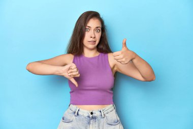 Mor bluzlu, mavi arka planda, başparmak yukarı ve baş parmak aşağı, zor bir seçim konsepti olan şık bir genç kadın.