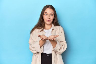 Mavi arka planda palto giymiş şık genç bir kadın avuçlarıyla bir şey tutuyor ve kameraya sunuyor..