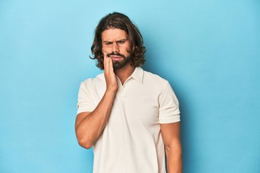 Beyaz polo giyen uzun saçlı bir adam, mavi stüdyo, güçlü bir diş ağrısı, azıdişi ağrısı var..
