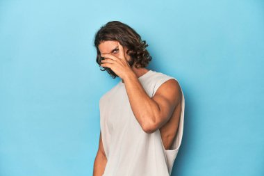 Askılı tişörtlü sakallı bir adam, mavi arka planda parmaklarıyla kameraya göz kırpıyor, yüzünü kapatmaktan utanıyor..