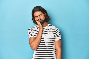 Çizgili gömlekli sakallı adam, mavi arka plan, sıkılmış, yorgun ve dinlenmeye ihtiyacı var..