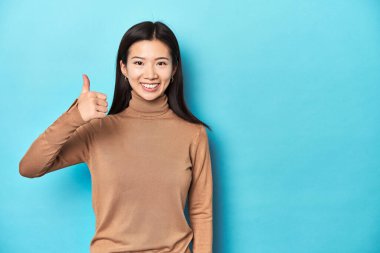 Kahverengi balıkçı yakalı genç Asyalı kadın, gülümsüyor ve başparmağını kaldırıyor.