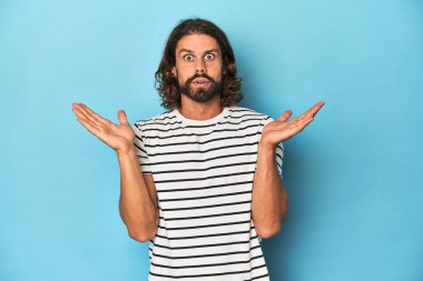 Çizgili gömlekli sakallı adam, mavi arka plan şaşırdı ve şok oldu..