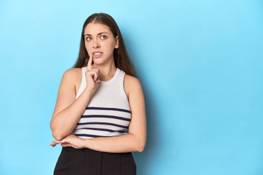 Çizgili bluzlu bir kadın mavi bir stüdyoda poz veriyor şüpheli ve şüpheci bir ifadeyle yana bakıyor..