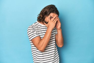 Çizgili gömlekli sakallı adam, mavi arka plan parmaklarının arasından gözünü kırp korkmuş ve gergin..