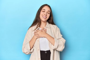 Mavi arka planda tişörtlü şık bir genç kadın. Gülüyor, ellerini kalbe koyuyor, mutluluk kavramını koruyor..