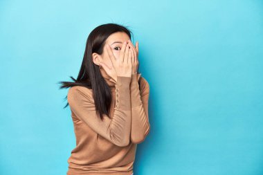 Kahverengi balıkçı yakalı genç Asyalı kadın, parmaklarının arasından korkmuş ve gergin bir şekilde göz kırp..