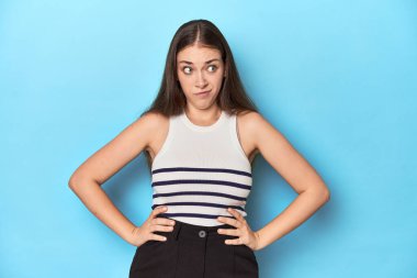 Çizgili bluzlu bir kadın mavi bir stüdyoda poz veriyor kafası karışık, kararsız ve kendinden emin değil..