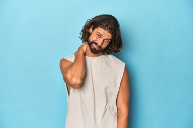 Askılı tişörtlü sakallı bir adam, mavi arka plan, sabit yaşam tarzından dolayı boyun ağrısı çekiyor..