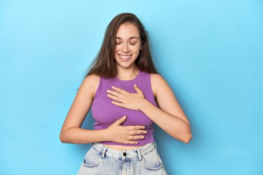 Mavi arka planda mor bluzlu şık bir kadın mutlu bir şekilde gülüyor ve ellerini karnının üstünde tutarak eğleniyor..