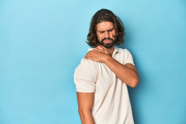 Beyaz polo giyen uzun saçlı bir adam, mavi stüdyoda omuz ağrısı çekiyor..