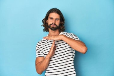 Çizgili gömlekli sakallı bir adam, mavi arka planda bir mola hareketi gösteriyor..