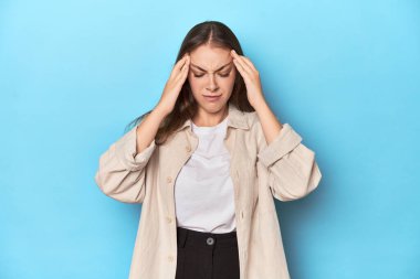 Mavi arka planda tişörtlü şık genç bir kadın şakaklara dokunuyor ve baş ağrısı çekiyor..