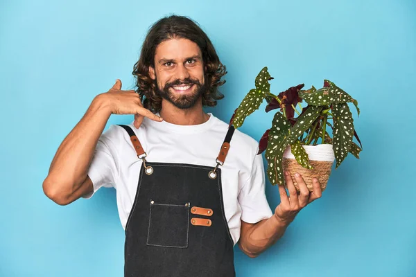 指で携帯電話のジェスチャーを示す植物を持っているひげを持つ長髪の庭師 — ストック写真