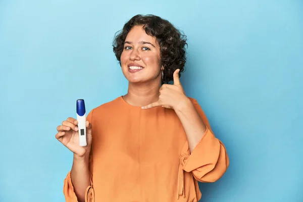 Young woman holding pregnancy test, studio background showing a mobile phone call gesture with fingers.