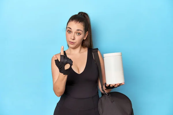 年轻女人拿着蛋白质瓶 站在运动的环境中 用手指指着你 好像邀请你走近些 — 图库照片
