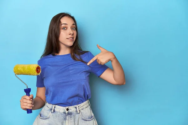 一个年轻的女人 带着黄色的彩绘滚筒 蓝色背景的人手指着衬衫的复制品空间 自豪而自信 — 图库照片