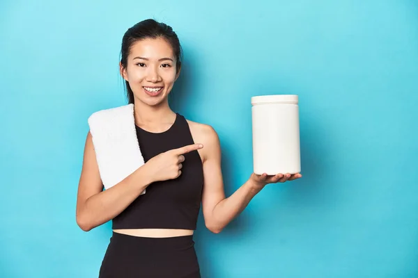 年轻的亚洲女运动员 带着蛋白质瓶 带着健身房的设备 微笑着指着旁边 在空白的空间里展示着一些东西 — 图库照片